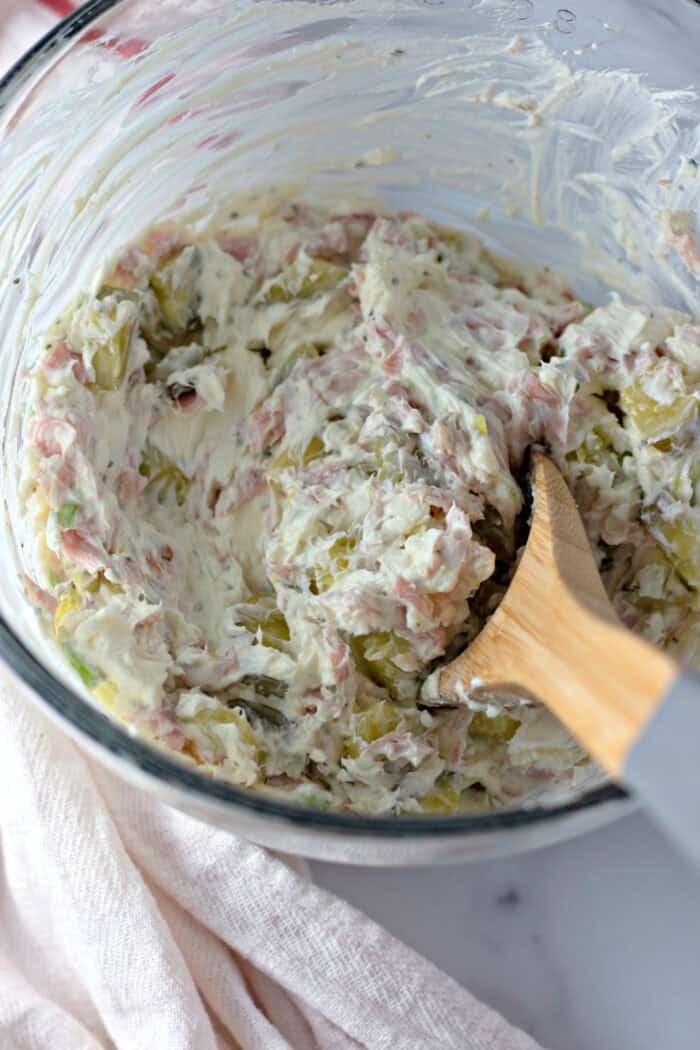 A wooden spoon stirring diced pickles, ham, and ranch seasoning into cream cheese