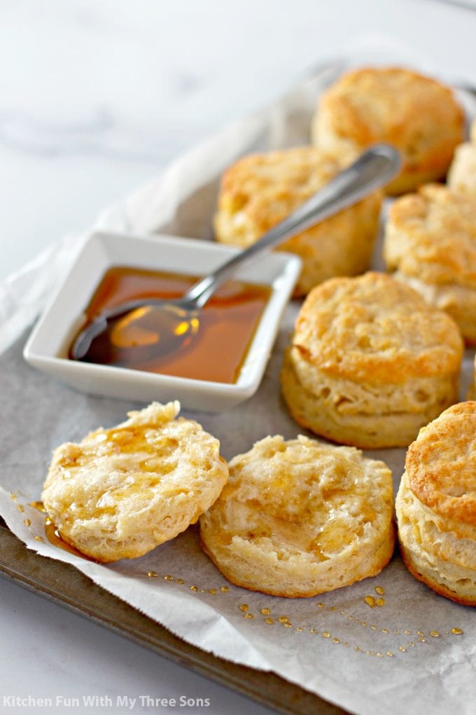 Honey Butter Biscuits on a cookie sheet with parchment paper and a bowl of honey