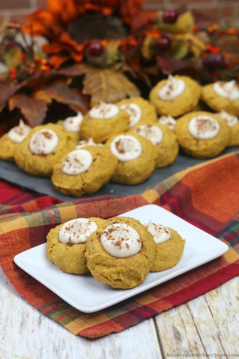 Three pumpkin thumbprint cookies on a plate in front of a tray of cookies