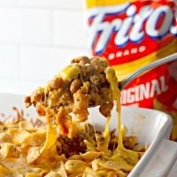 A casserole filled with Easy Frito Pie. A spoon is lifting out a scoop of the Frito Pie, and the cheese is melting and stretching from the casserole up to the spoon. A bag of Fritos is visible in the background of the shot.
