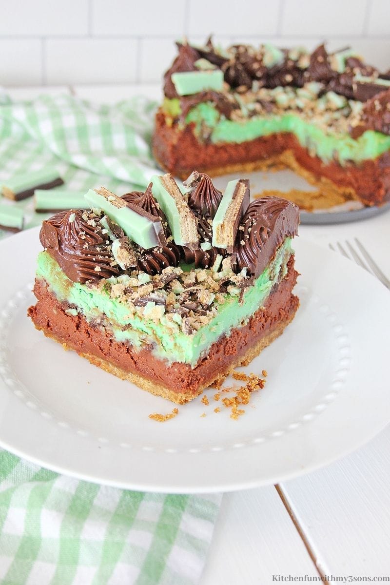 A slice of Kit Kat Chocolate Mint Cheesecake on a serving plate with a green patterned cloth around it.