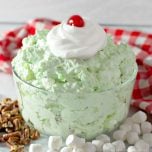 Watergate Salad in a clear bowl topped with whipped cream and a cherry, next to mini marshmallows and chopped pecans.