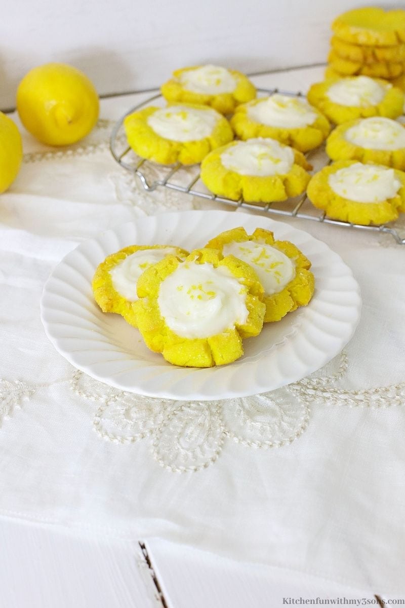 Lemon sugar cookies on a serving plate with full lemons behind it and extra cookies on a wire rack.