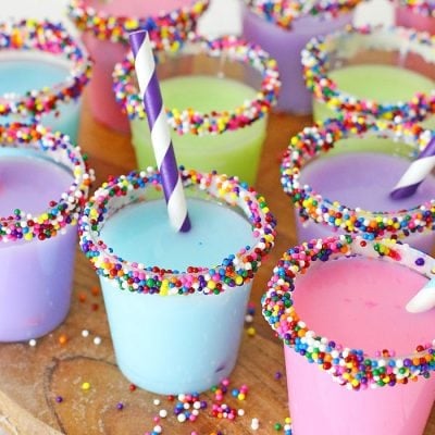 Assorted colorful birthday cake shots rimmed with frosting and rainbow sprinkles.