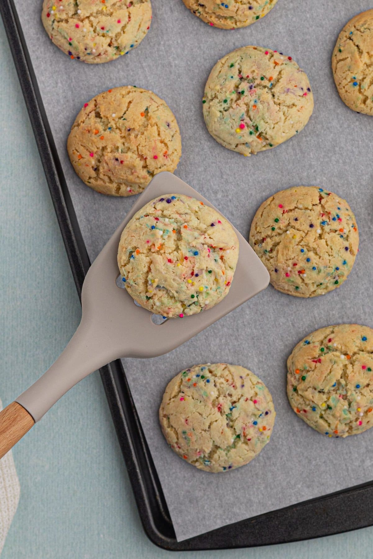 A parchment-lined baking sheet with baked sprinkle cookies. One cookie is being lifted out on a silicone spatula.