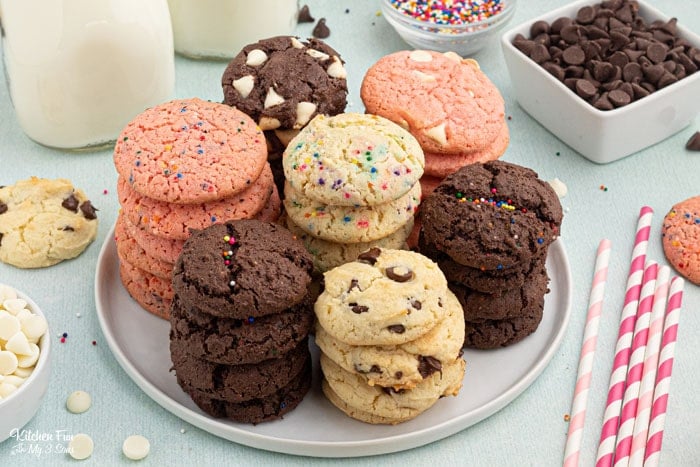 A plate of seven different kinds of cookies, stacked in seven columns. Dishes of chocolate chips, white chocolate chip, and sprinkles are arranged around the table, along with party straws and mason jars of milk.