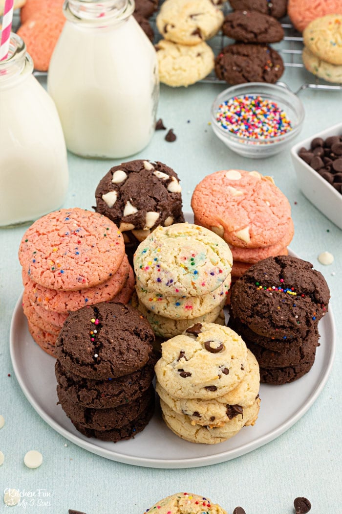 A platter of different kinds of cookies, some chocolate, some strawberry, and some vanilla. Glasses of milk and small dishes of toppings in the background.