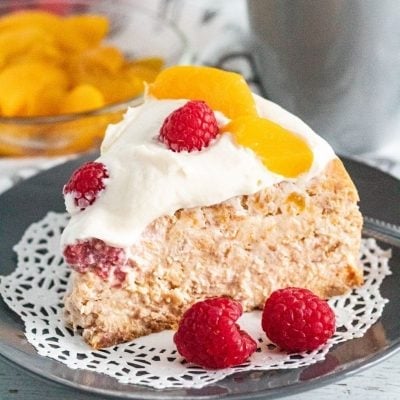A slice of cheesecake surround by raspberries and peaches on a gray plate and white doily.