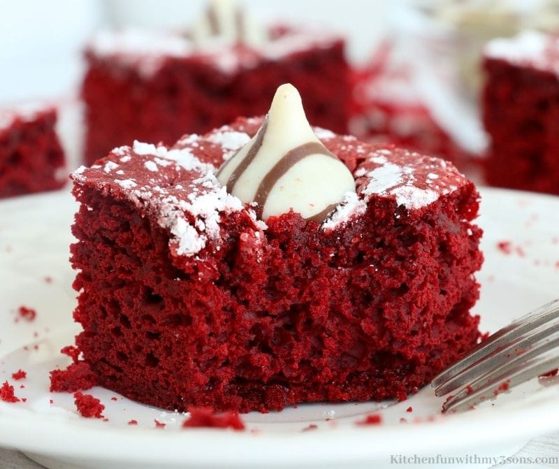 A square slice of Red Velvet Cake topped with powdered sugar and a kiss on a white plate