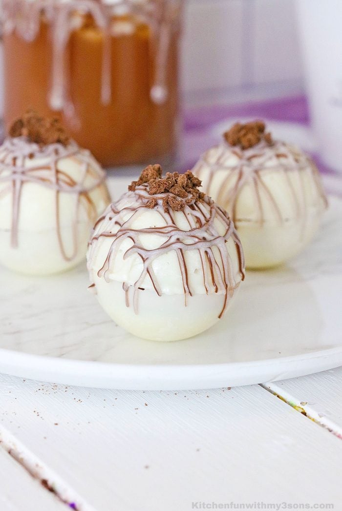 White hot cocoa bombs on a white plate, drizzled with caramel and crushed chocolate.
