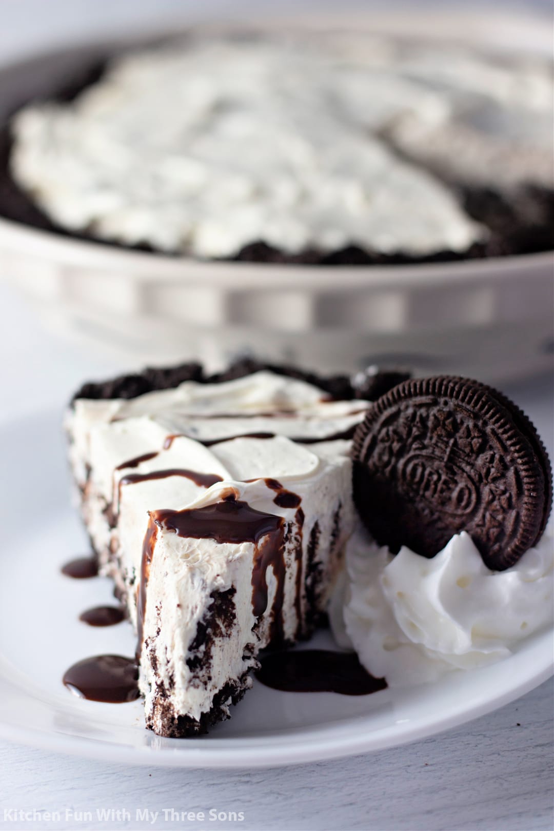 A slice of no-bake Oreo pie on a plate, drizzled with chocolate sauce next to whipped cream garnished with an Oreo cookie, with the rest of the pie in the background.