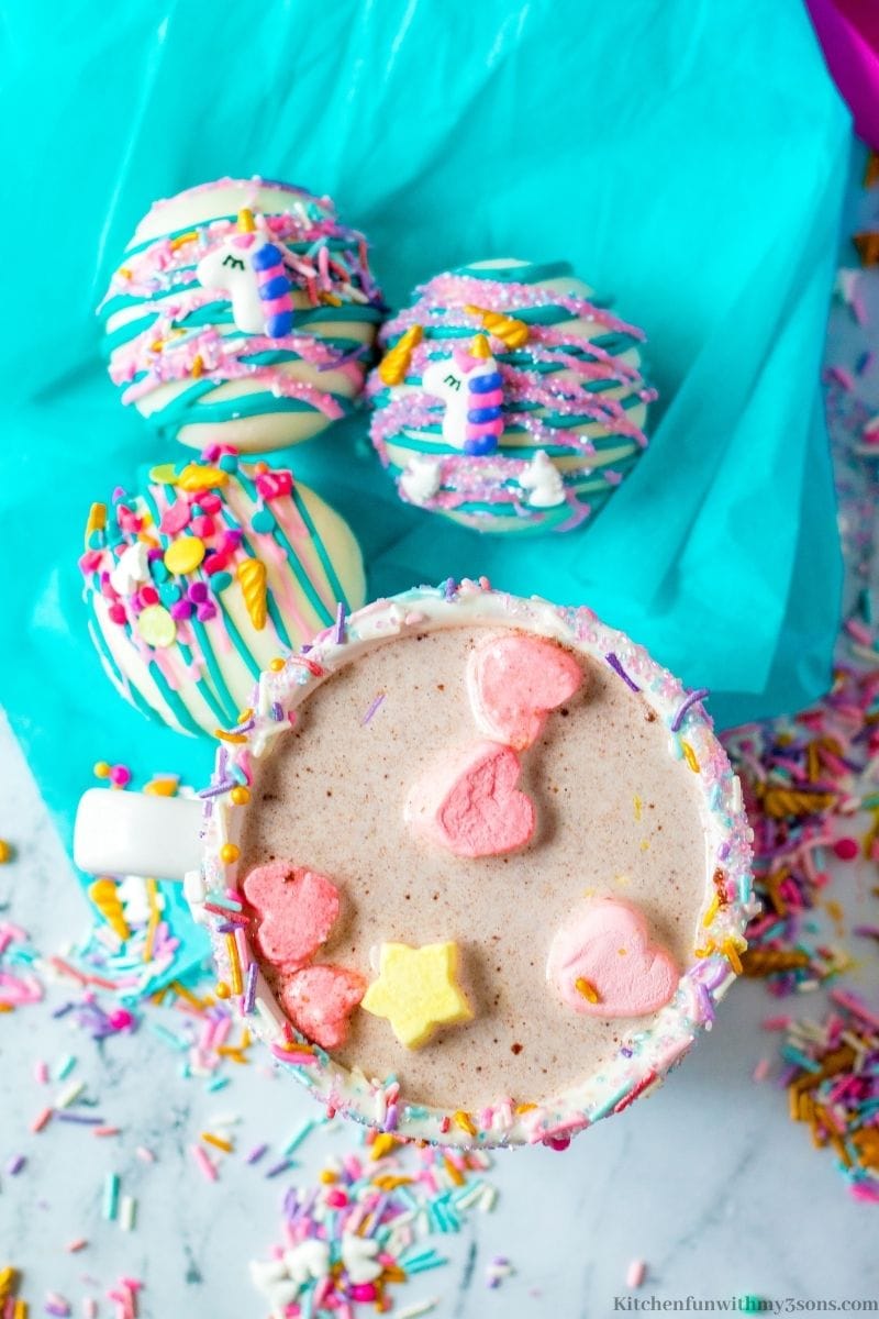 A melted cup of hot cocoa with three Unicorn Hot Chocolate Bombs next to it.