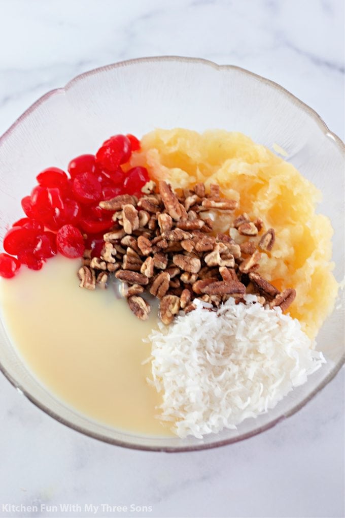sweetened condensed milk, coconut, maraschino cherries, pecans, and pineapple in a clear bowl.