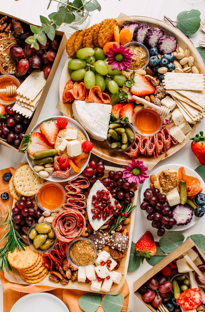 Overhead view of various charcuterie boards with meats, cheese, crackers, fruits, honey, pickles, and more