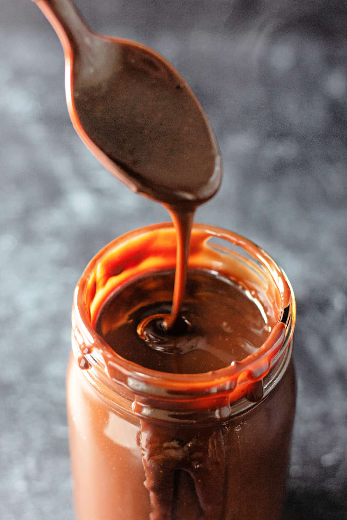 Ready in 5 minutes with just 6 basic ingredients, this Hot Fudge Sauce is rich, decadent, and incredibly chocolatey. A spoonful of hot fudge above a jar with the fudge drizzling into the jar
