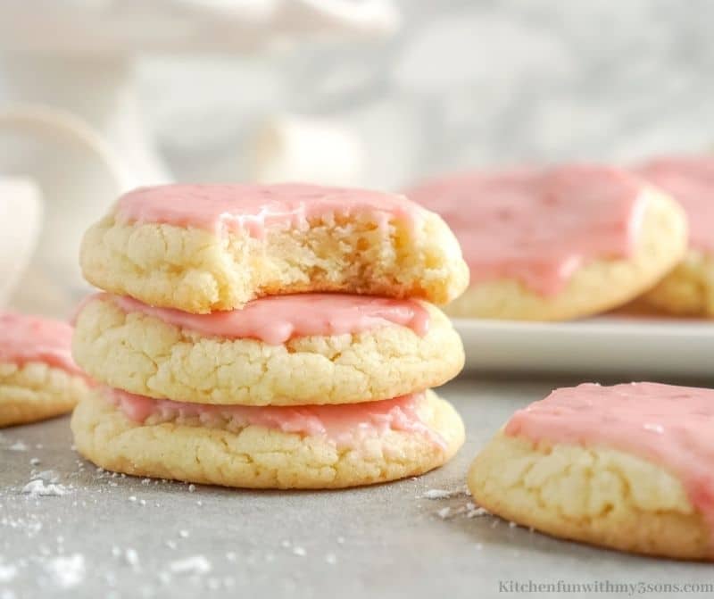 Three strawberry sugar cookies stacked, with a bite missing from the top cookie and more cookies in the background.