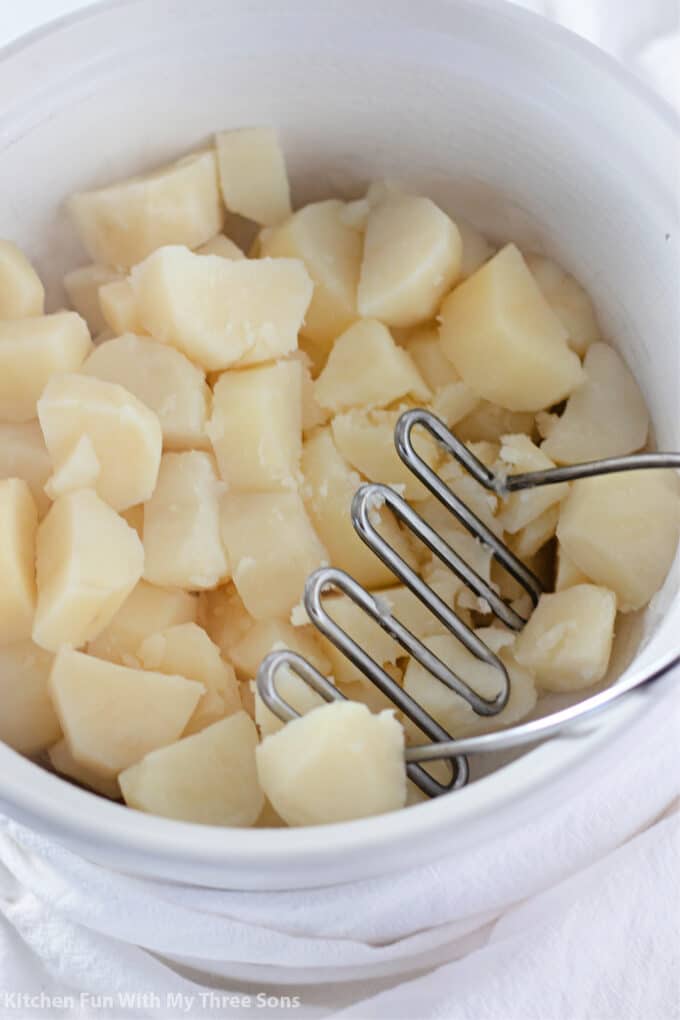 boiled potato chunks in a white mixing bowl with a potato masher.