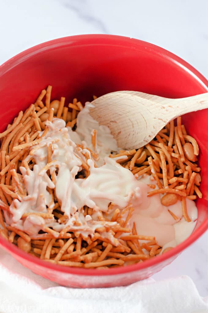 mixing together the white chocolate, peanuts, and chow mein noodles in a red bowl with a wood spoon.