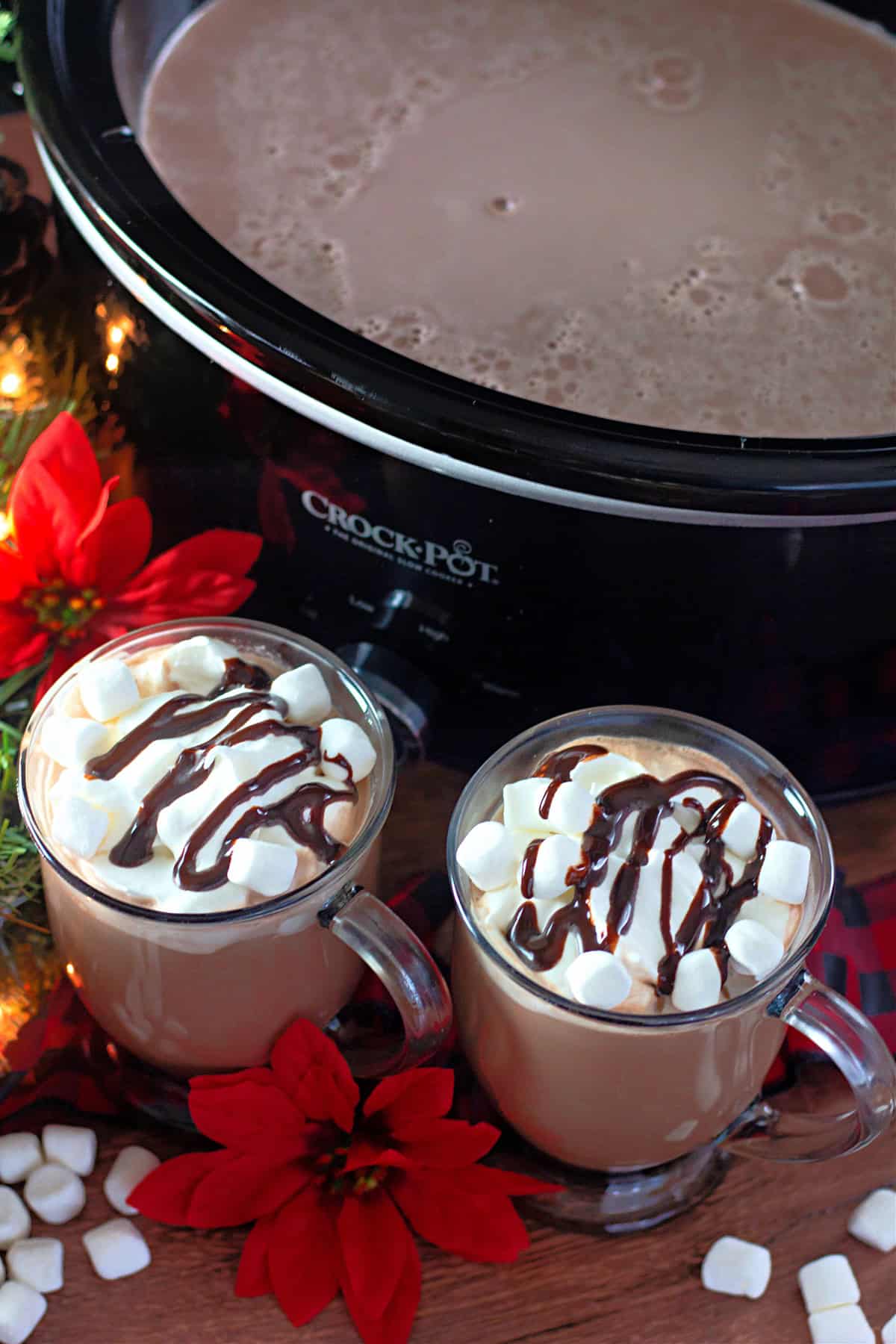 Two mugs of hot chocolate topped with whipped cream and chocolate in front of the slow cooker