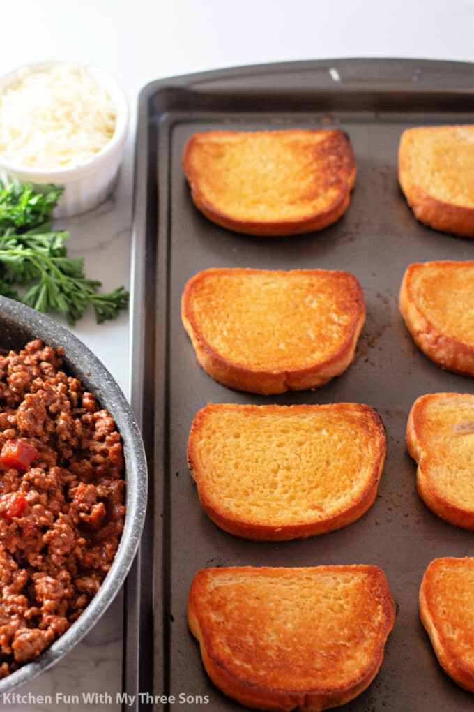 garlic toast on a cookie sheet next to meat sauce, parsley, and mozzarella cheese.