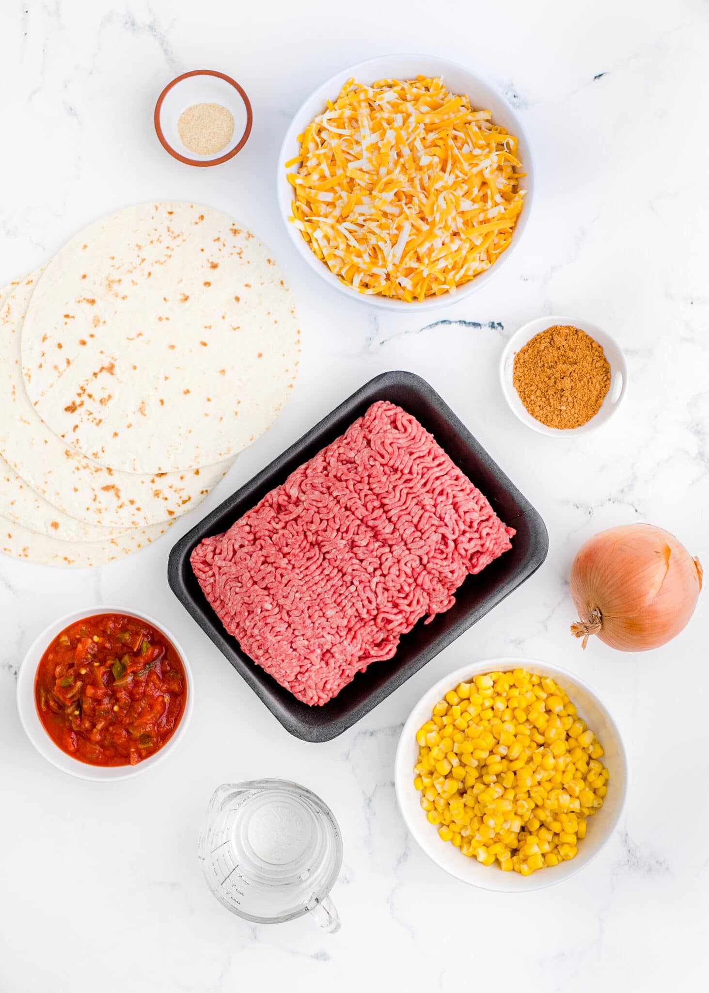 Clockwise from top: Garlic powder, cheese, taco seasoning, onion, corn, water, salsa, tortillas. Center: A package of ground beef.