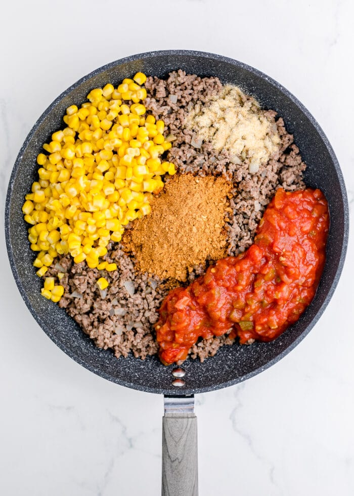 A skillet full of browned ground beef with corn, seasonings, and salsa dumped in.