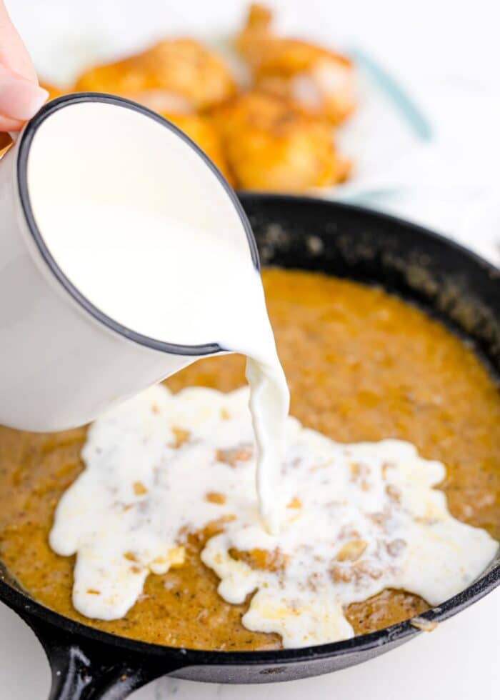 Milk being poured from an enamel jug into a cast-iron skillet full of chicken gravy.
