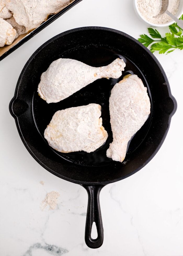 A cast-iron skillet with three flour-dredged chicken pieces.