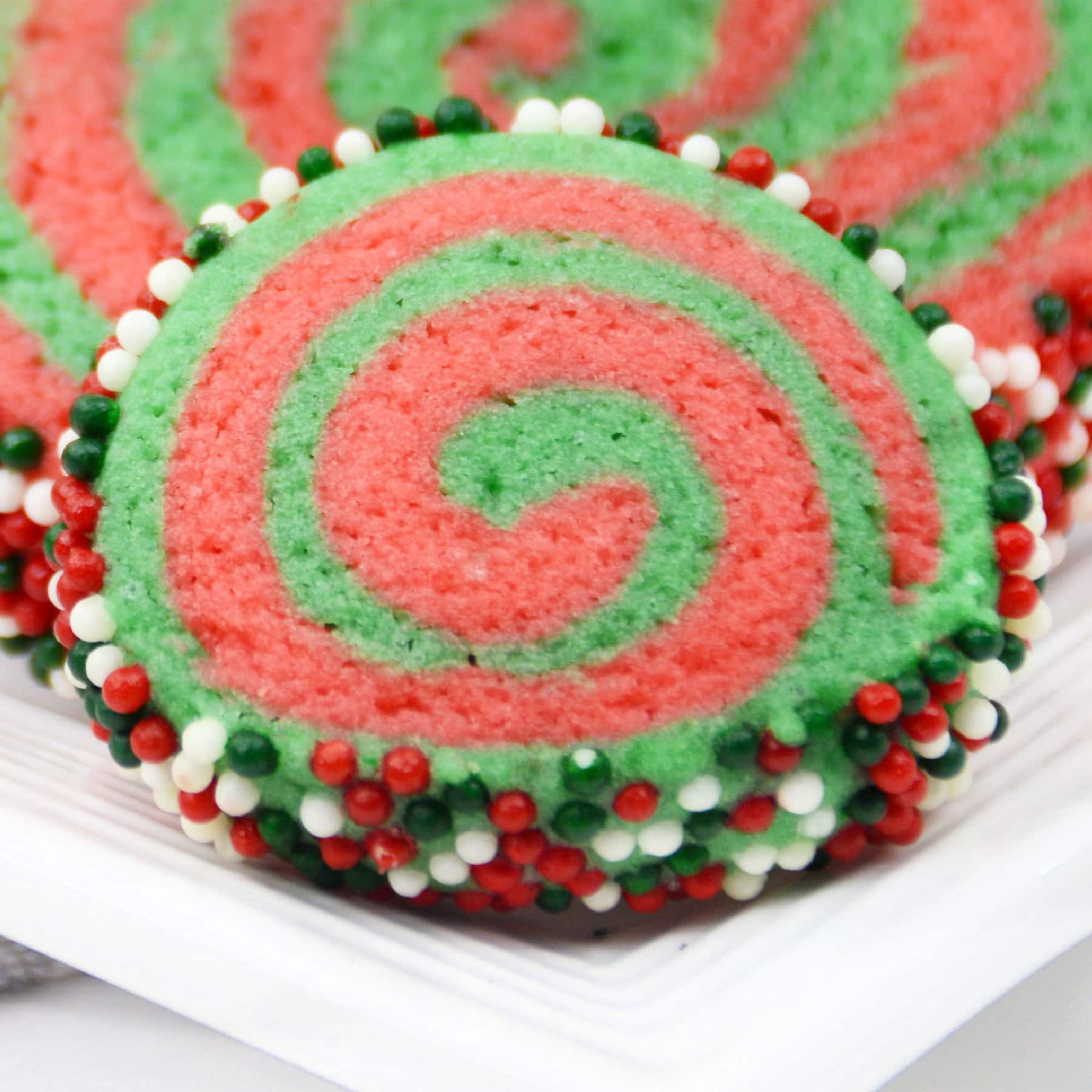 Close up of red and green christmas pinwheel cookie rolled in sprinkles on a white plate.