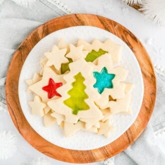 A plate full of stained glass cookies on a wooden circular board, surrounded by white Christmas decorations.