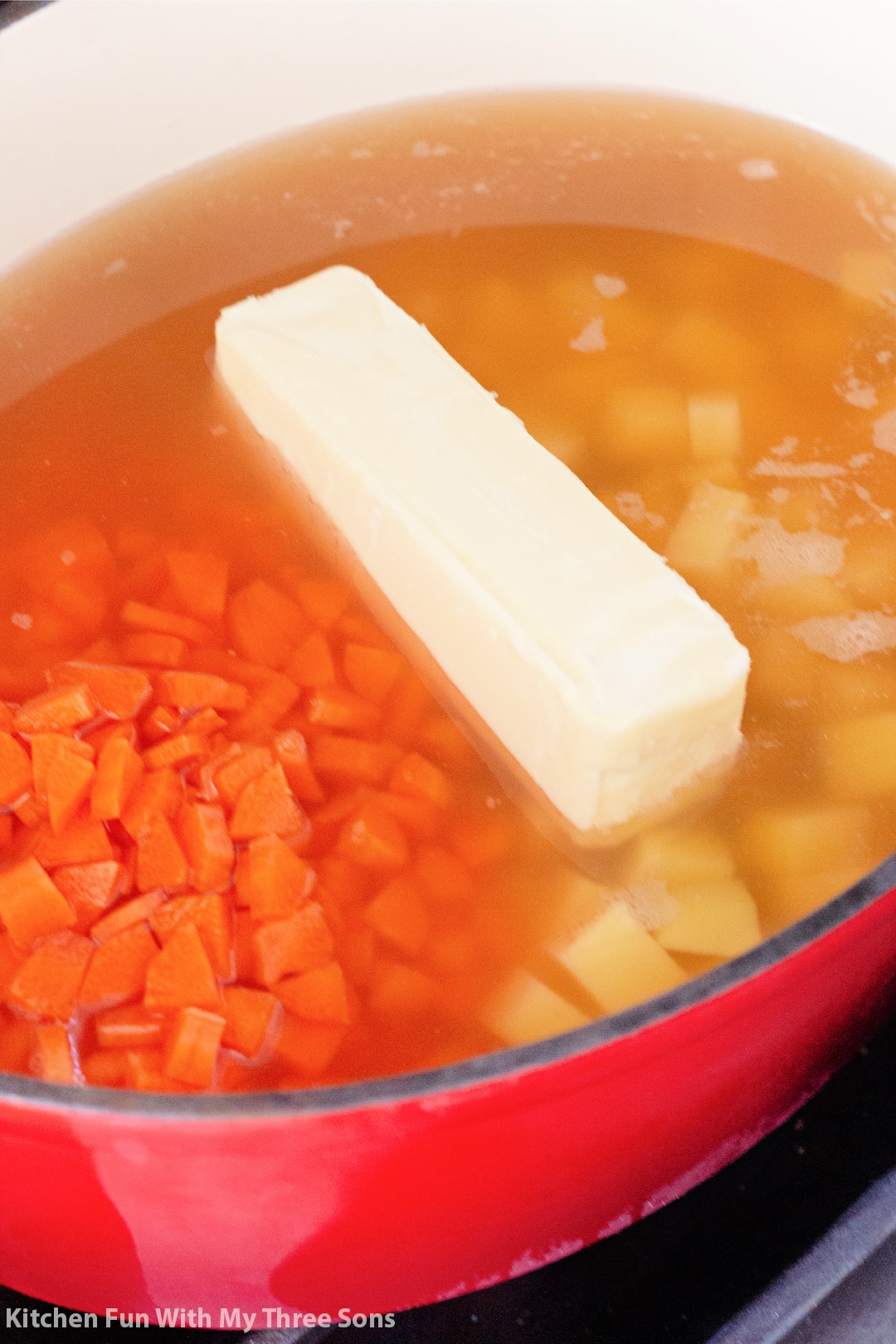 A stick of butter melting into a pot full of chicken broth, diced carrots and cubed potatoes