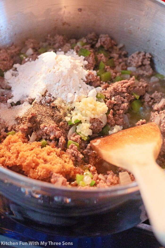 ground beef, onion, garlic, green bell pepper, brown sugar, salt, pepper, and flour in a large pot with a wooden spoon.