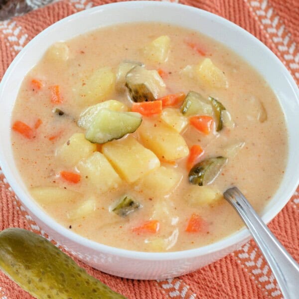 A bowl of soup with chunks of pickle, carrot and potato and a pickle next to it