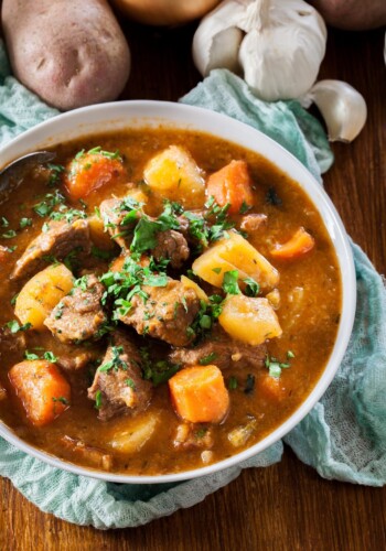 A bowl of beef stew on top of a wooden table with potatoes and garlic behind it
