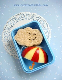 A sandwich cut into a cloud shape beside a halved apple decorated to look line an umbrella inside of a blue lunch box