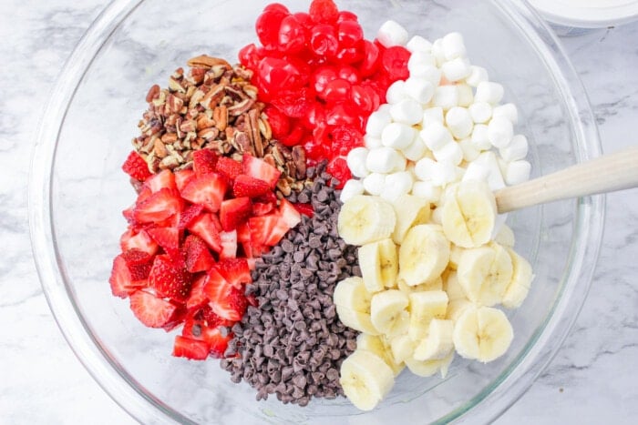 Bananas, marshmallows, chocolate chips, cherries, strawberries, and nuts in a glass bowl