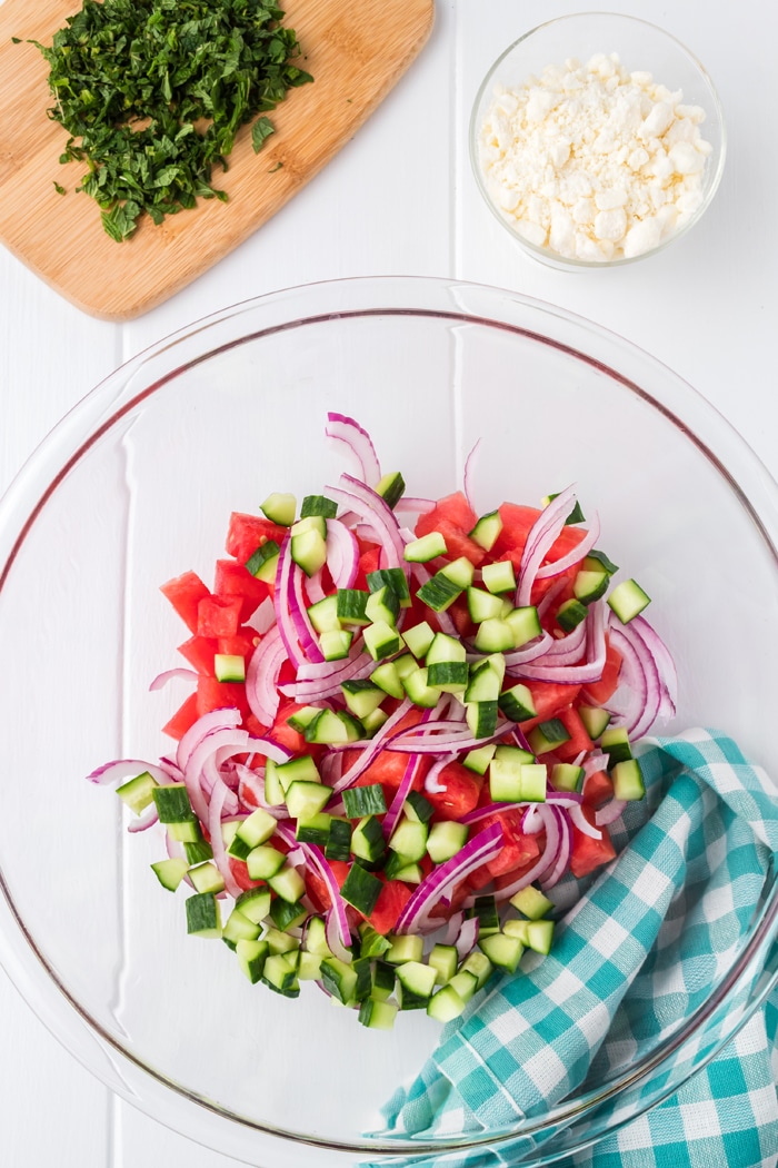 Overhead view of watermelon salad ingredients combined in a glass bowl, next to a bowl of feta and chopped basil on a cutting board.