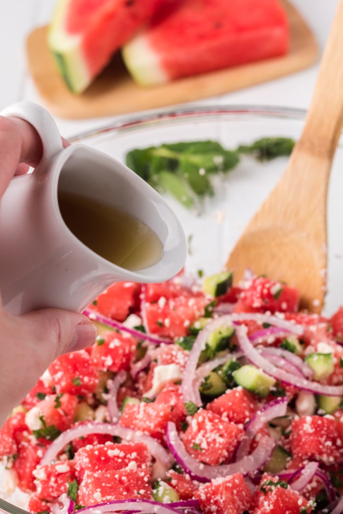 A hand pouring dressing over a watermelon salad from a small ceramic pourer, with watermelon slices on a cutting board in the background.