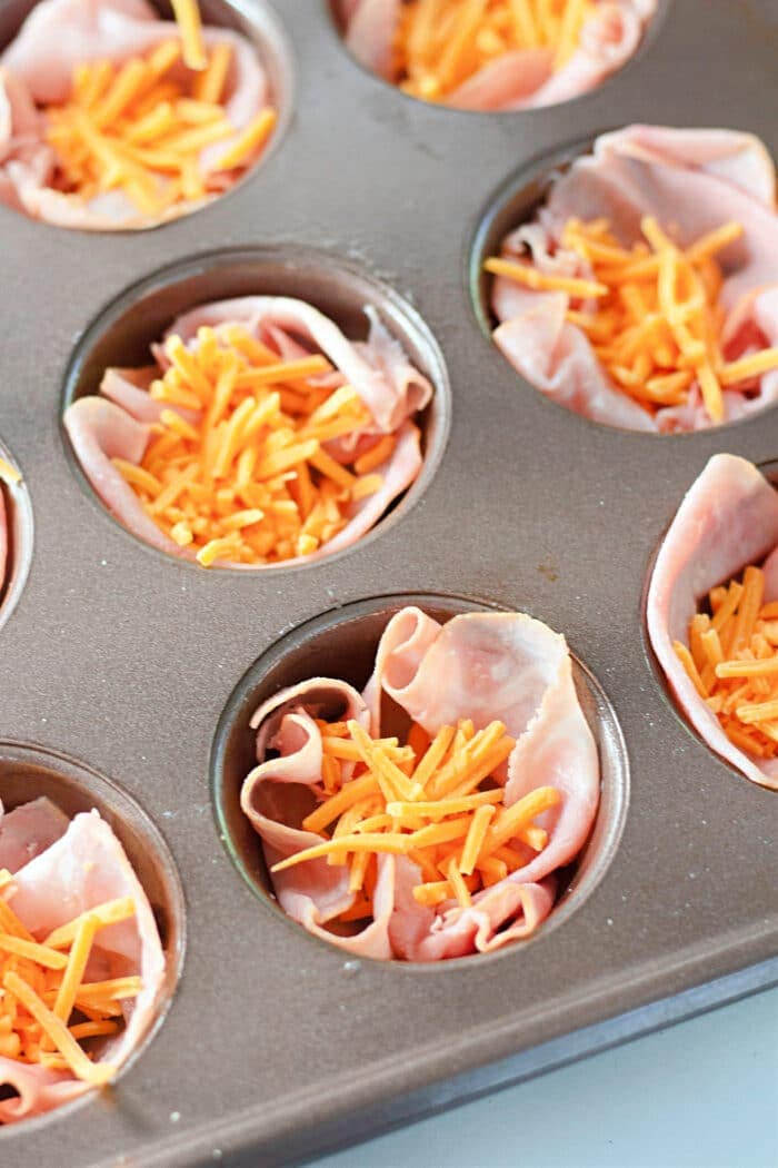 slices of ham and shredded cheese in a muffin tin.