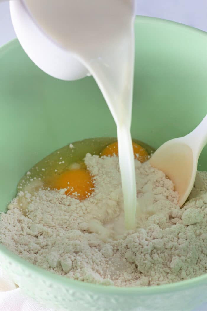 pouring milk into a green bowl of pancake mix and eggs.