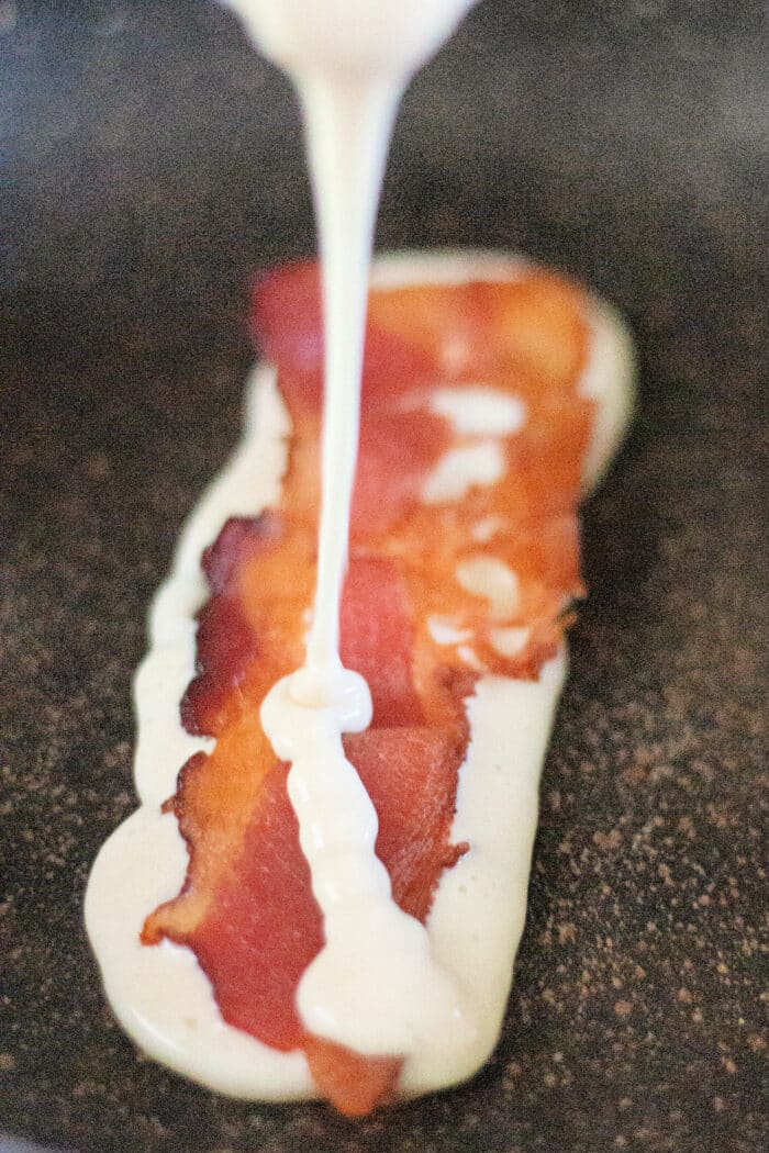 ladling pancake batter over a strip of bacon on a skillet.