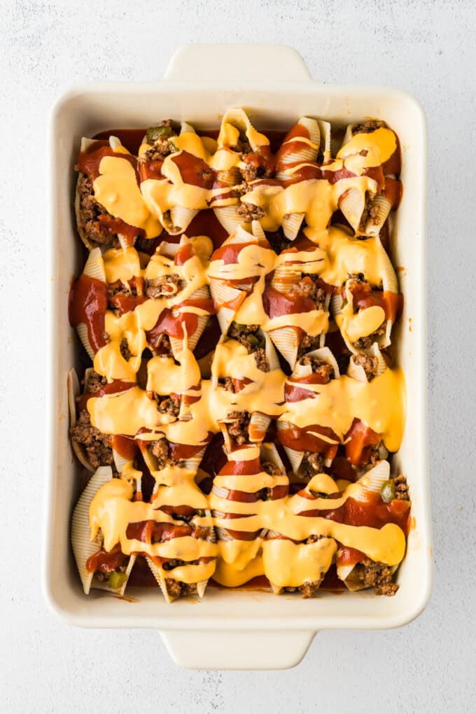 Jumbo shells are stuffed with seasoned ground beef and topped with queso, taco sauce, and shredded cheese to make these Taco Stuffed Shells. Taco sauce and queso drizzled over stuffed shells in a casserole dish