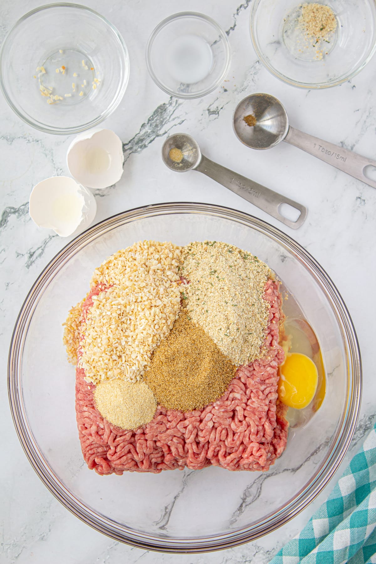 A bowl of ground beef with an egg cracked into it and seasonings piled on top.