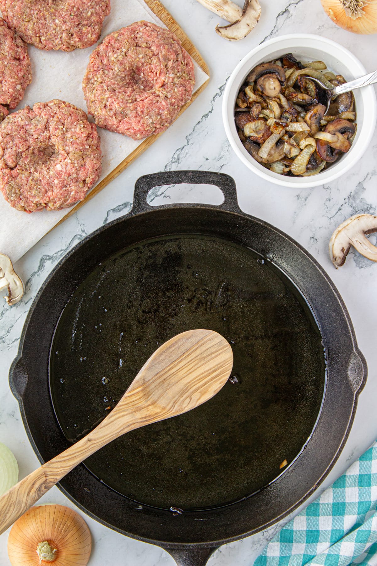 A greased pan next to four steak patties and a bowl of mushrooms and onions.