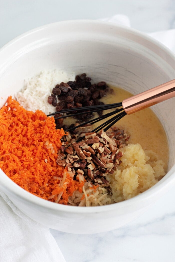 shredded carrot, coconut, pineapple, pecans, raisins, and eggs in a white mixing bowl with a whisk.