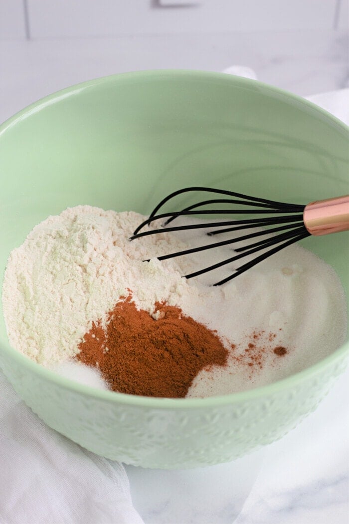 whisking together flour, sugar, baking soda, salt, and cinnamon in a mint green mixing bowl.