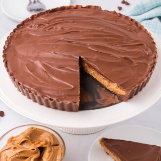 A Reese's Peanut Butter Cup Pie on a cake stand next to a slice of pie on a white plate, and a bowl of peanut butter.