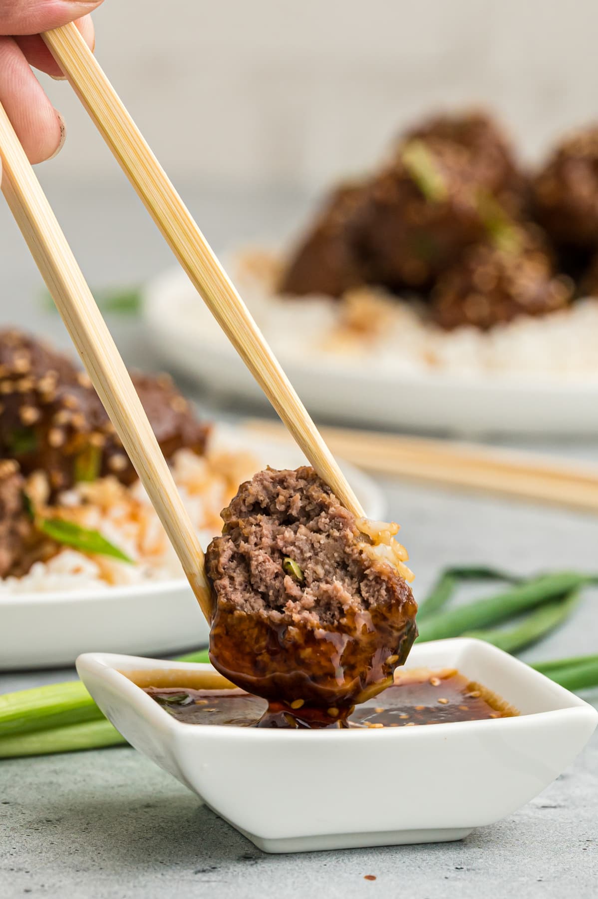 A meatball between two chopsticks being dipped into teriyaki sauce