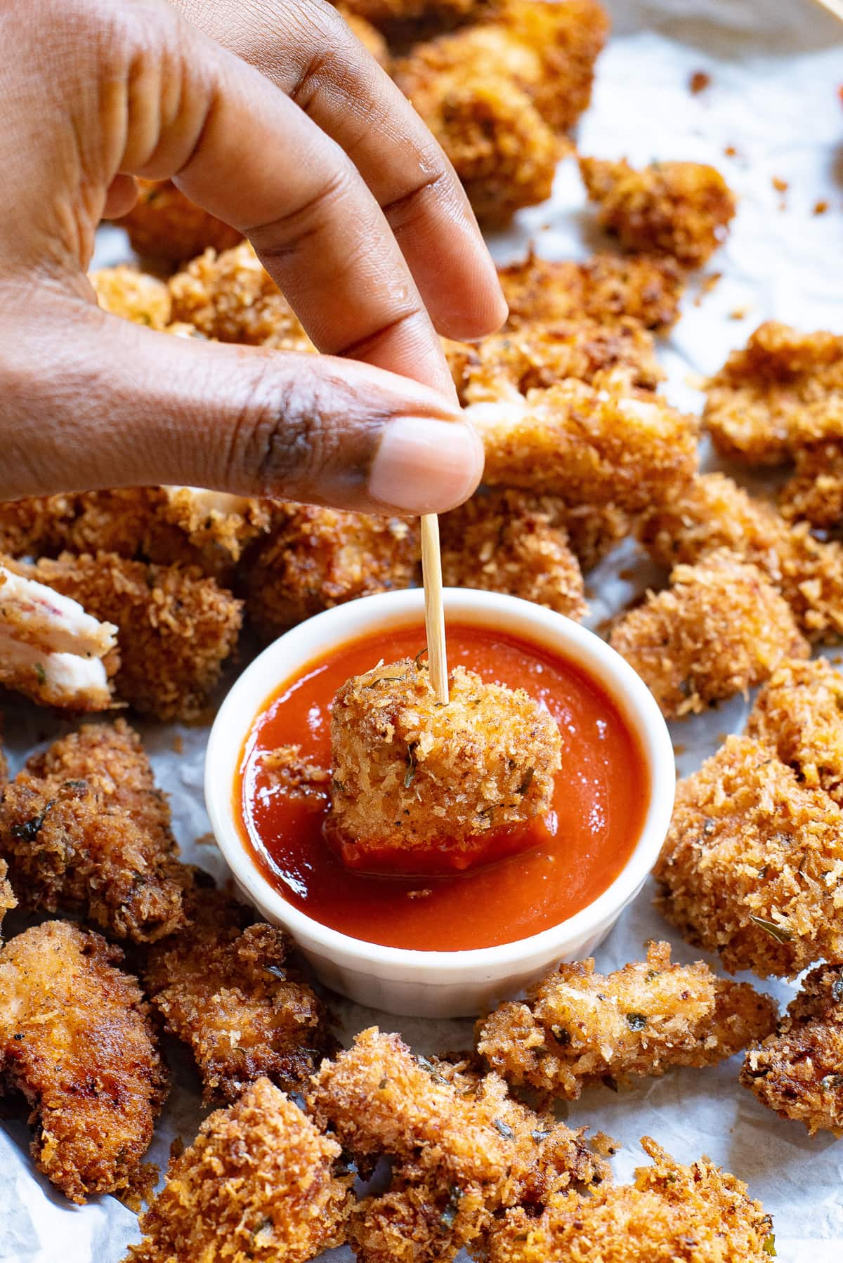 A hand holding a piece of popcorn chicken on a toothpick dipping it into ketchup