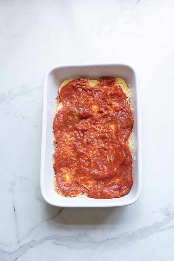 sauce on top of cheese for baked ravioli in white rectangle baking dish